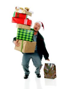 A man in a Santa hat attempting to balance a huge stack of wrapped Christmas gifts while reaching for another.